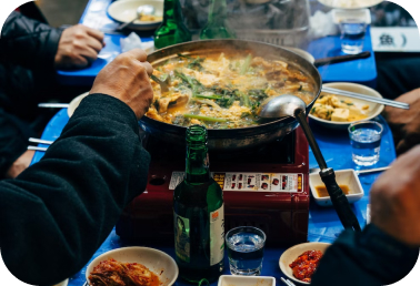 A group of people gathered around a boiling pot of food, each with a plate and drink in front of them.