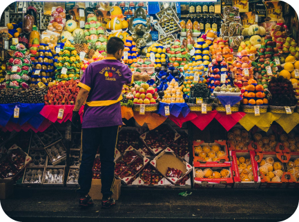 A person in a purple shirt stands in front of a fruit stand filled with various colorful fruits.